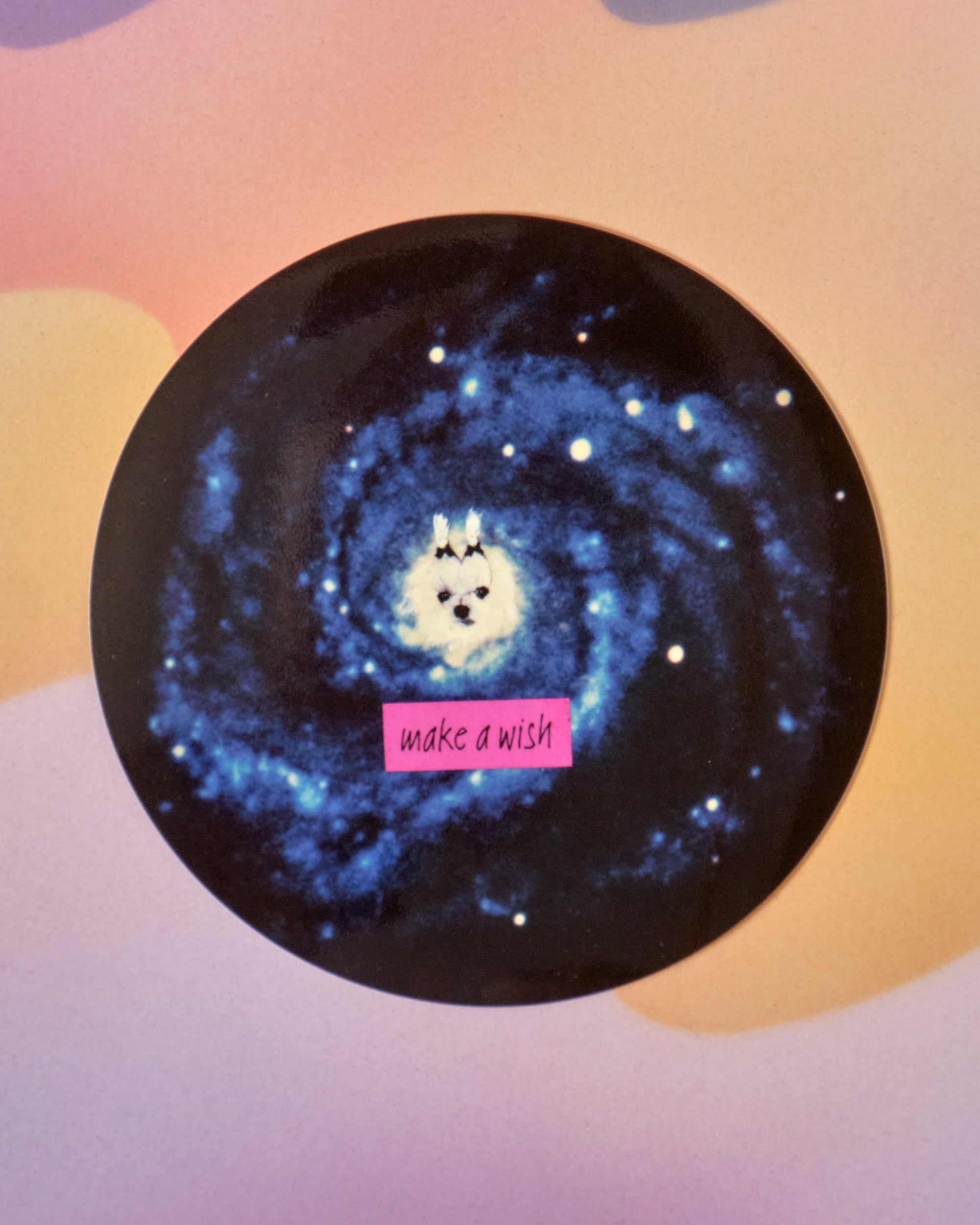 Circular sticker featuring white fluffy dog in blue galaxy spiral with pink 'make a wish' text