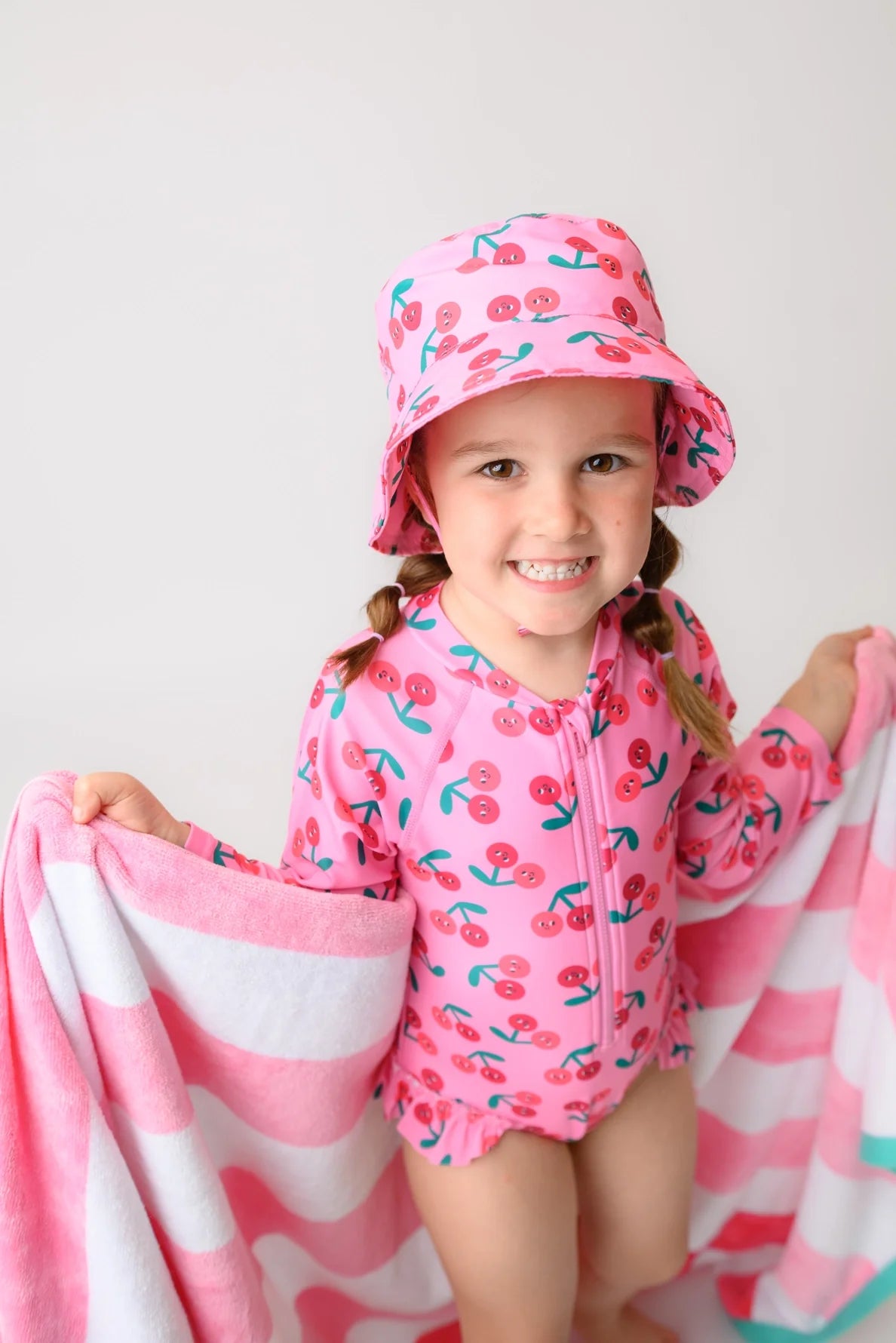 Young girl wearing pink cherry-print rash guard swimsuit and matching bucket hat holding striped towel
