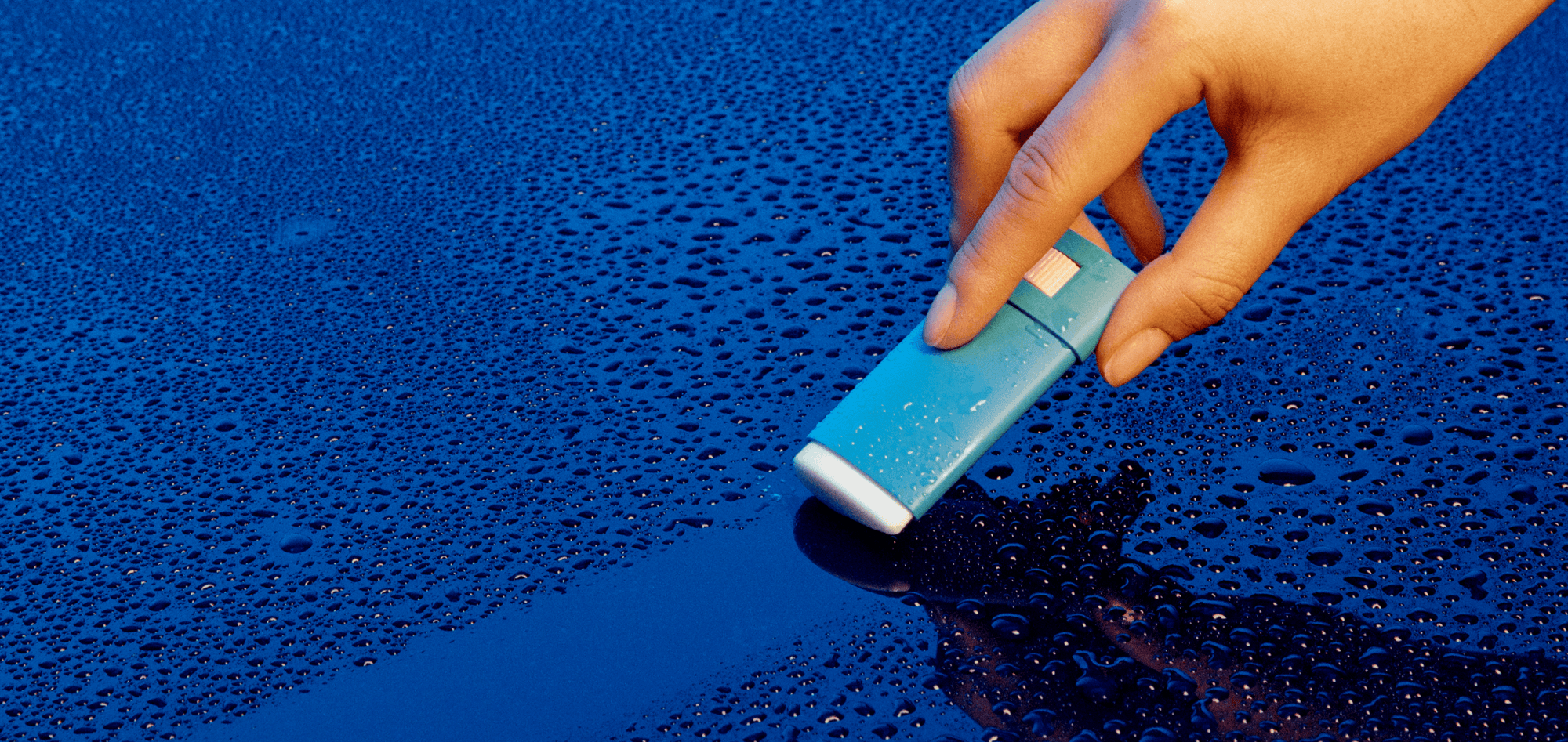 Hand holding turquoise Vacation Super Stick SPF 45 sunscreen on wet blue surface