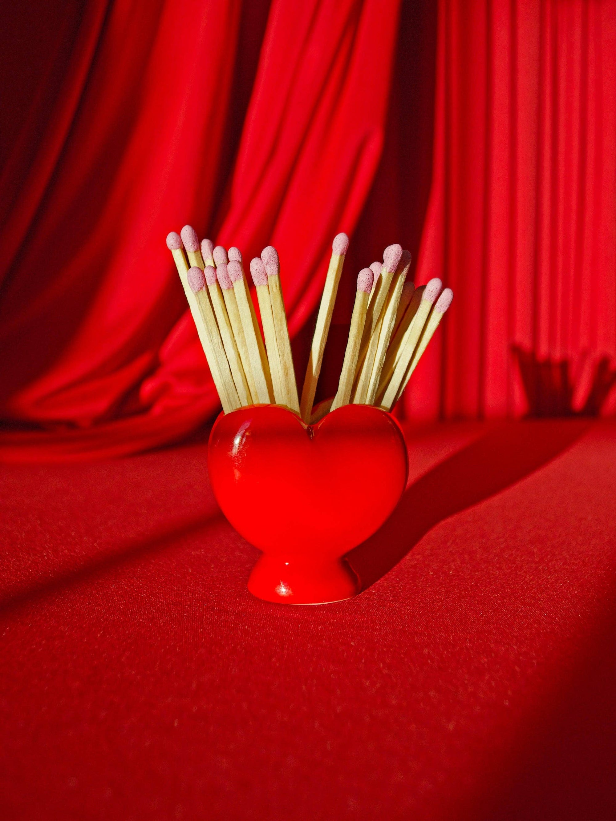 Red heart-shaped match striker holder with wooden matches against red fabric backdrop