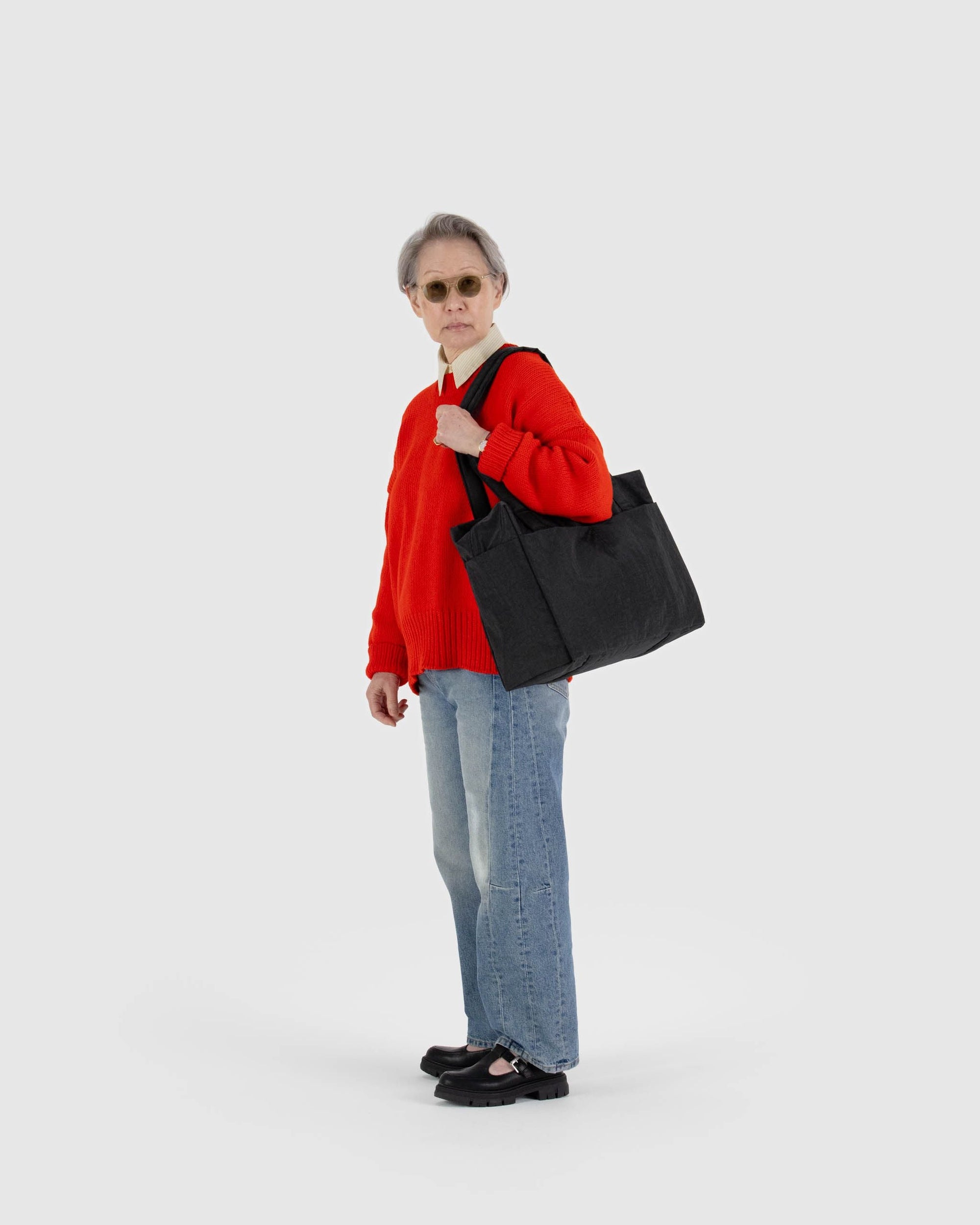 Person wearing red sweater and jeans carrying black Baggu Cloud Carry-On bag over shoulder