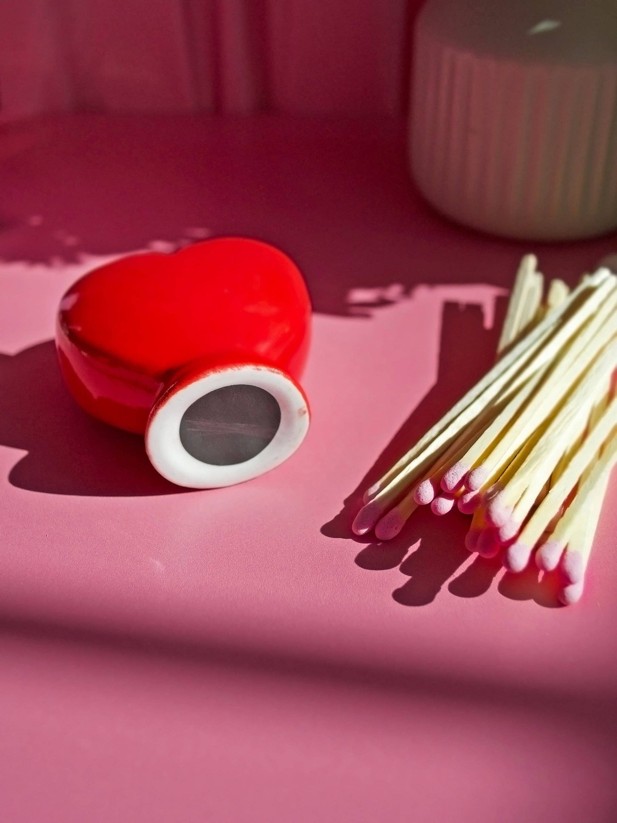 Red heart-shaped match striker with white rim next to wooden matches on pink surface