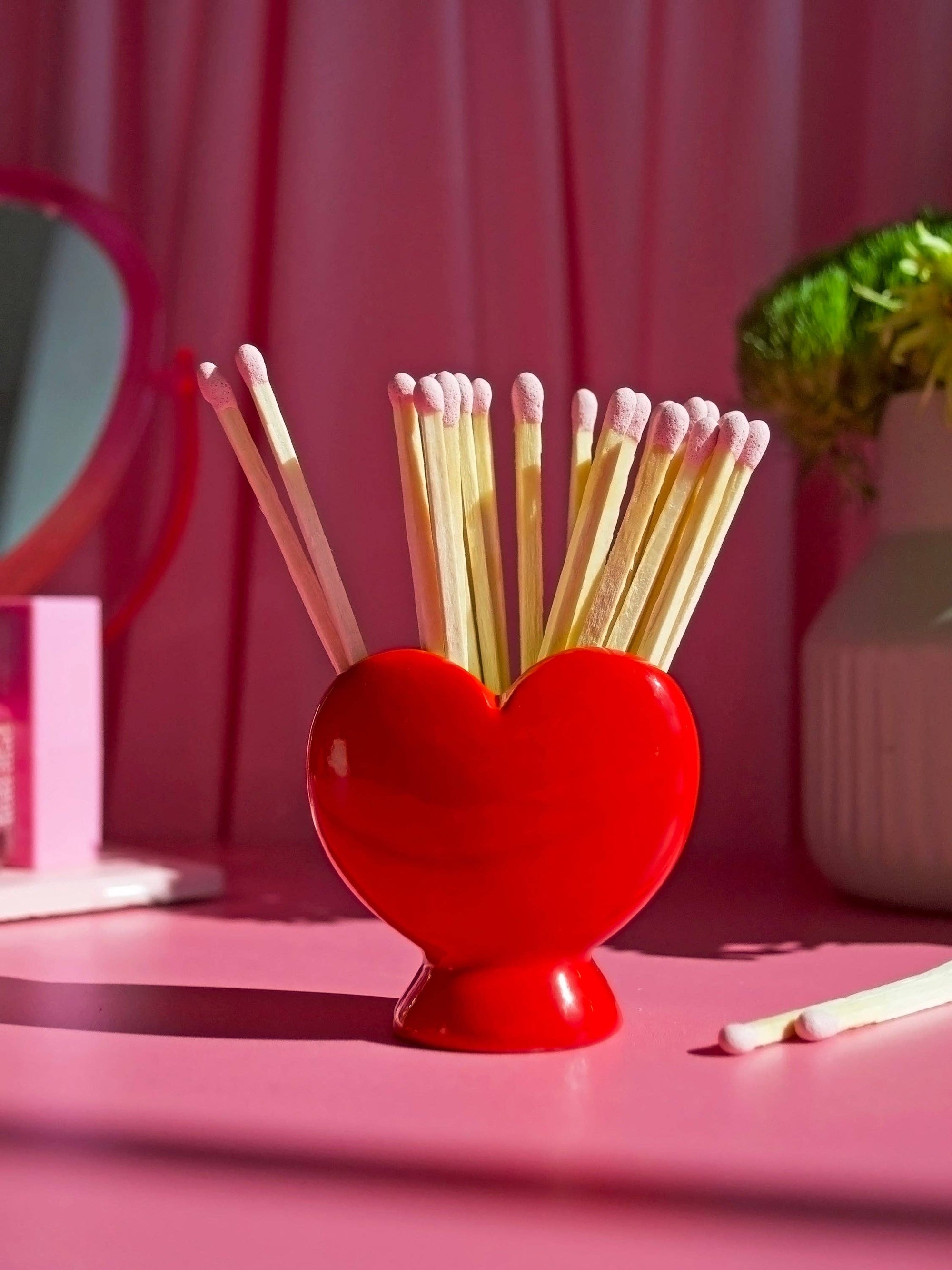 Red heart-shaped match striker holder with wooden matches against pink background