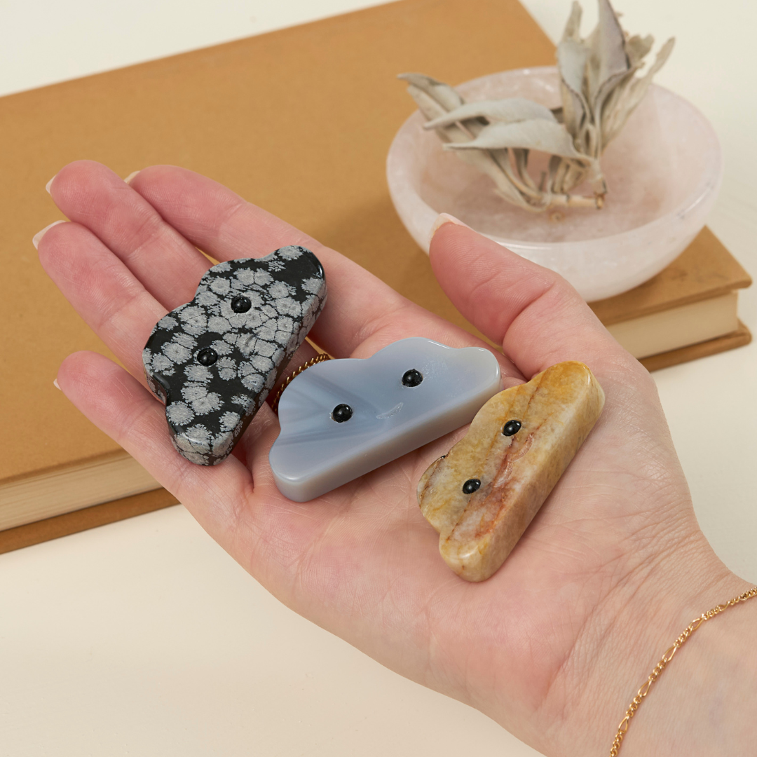 Hand holding three crystal cloud-shaped pet rocks in black, blue, and tan with dot eyes