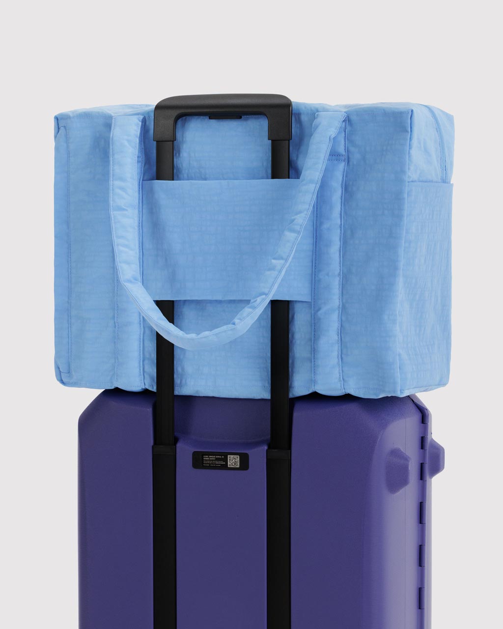 Light blue Baggu Cloud Carry-on tote bag resting on top of a purple hard-shell rolling suitcase