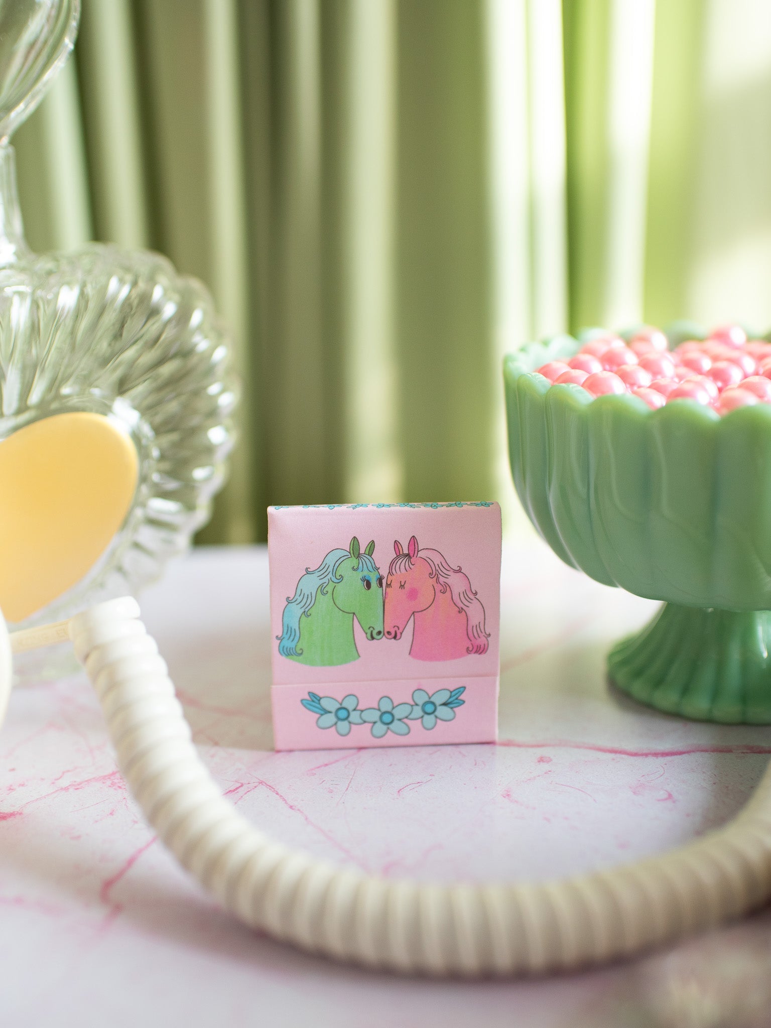 Pink matchbook with two cartoon horses nuzzling and floral design on white surface