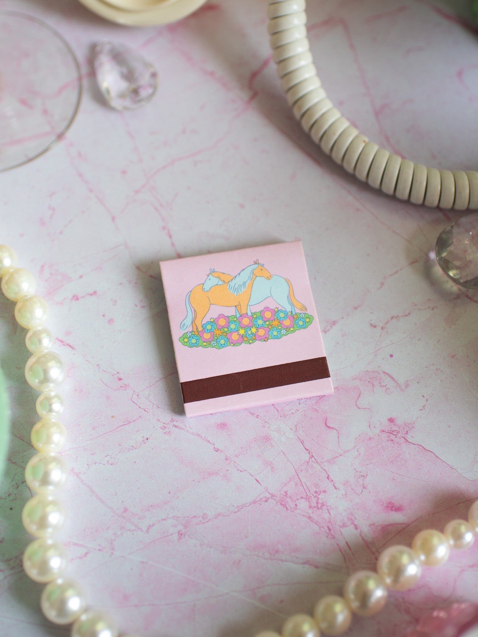 Pink matchbook with illustrated horses and flowers surrounded by pearl jewelry on marble surface