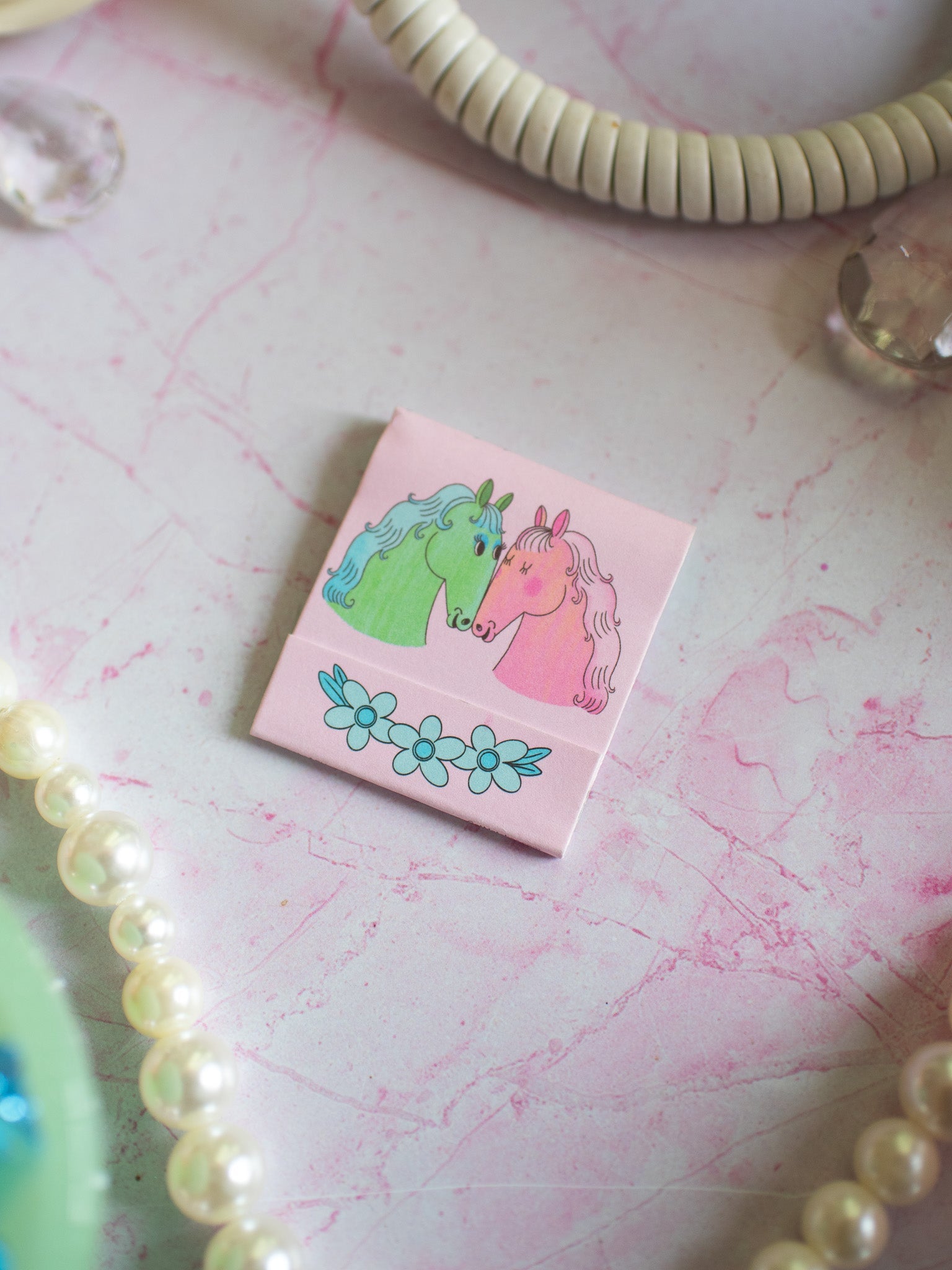 Pink matchbook with illustrated unicorn couple and blue flowers on marble surface with pearls