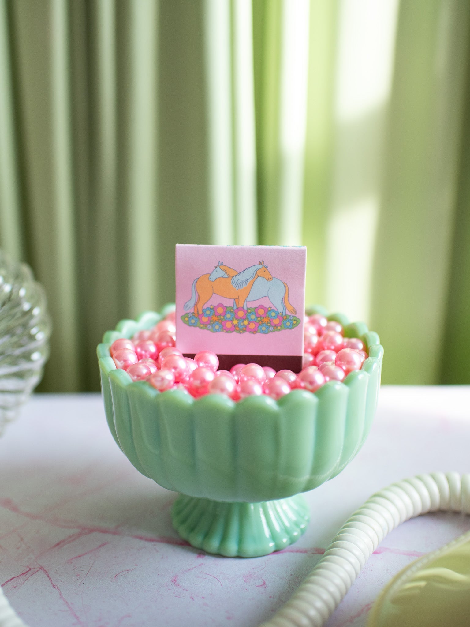 Pink horse-themed matchbook in mint green decorative bowl with pink candies on white table