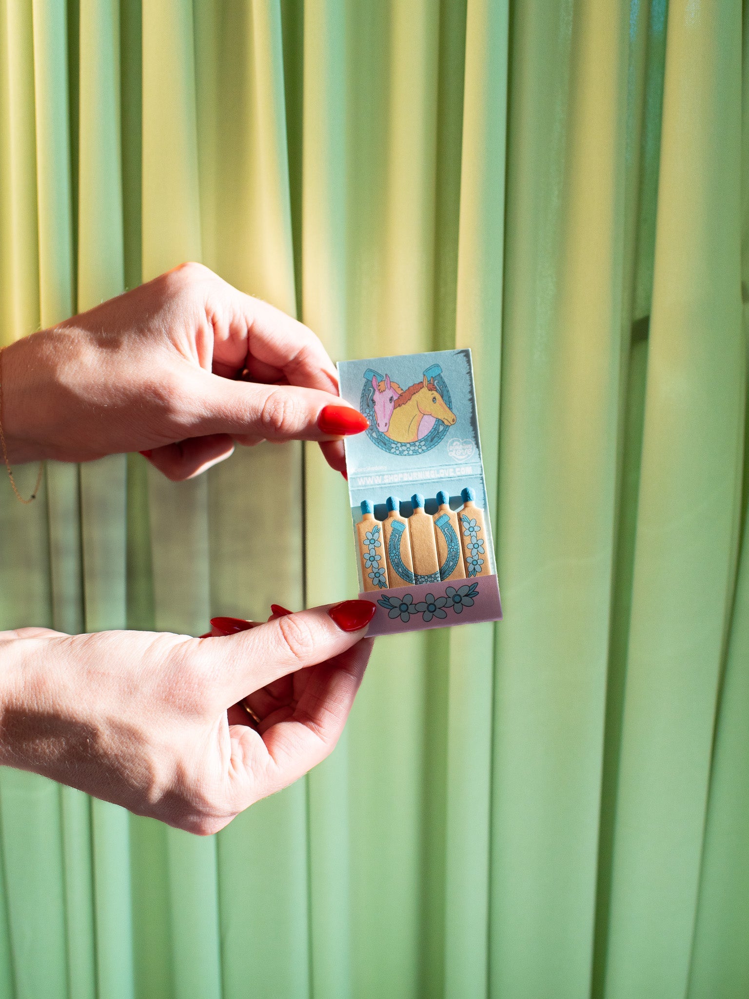 Hands with red nails holding colorful matchbook featuring horse illustration and decorative design