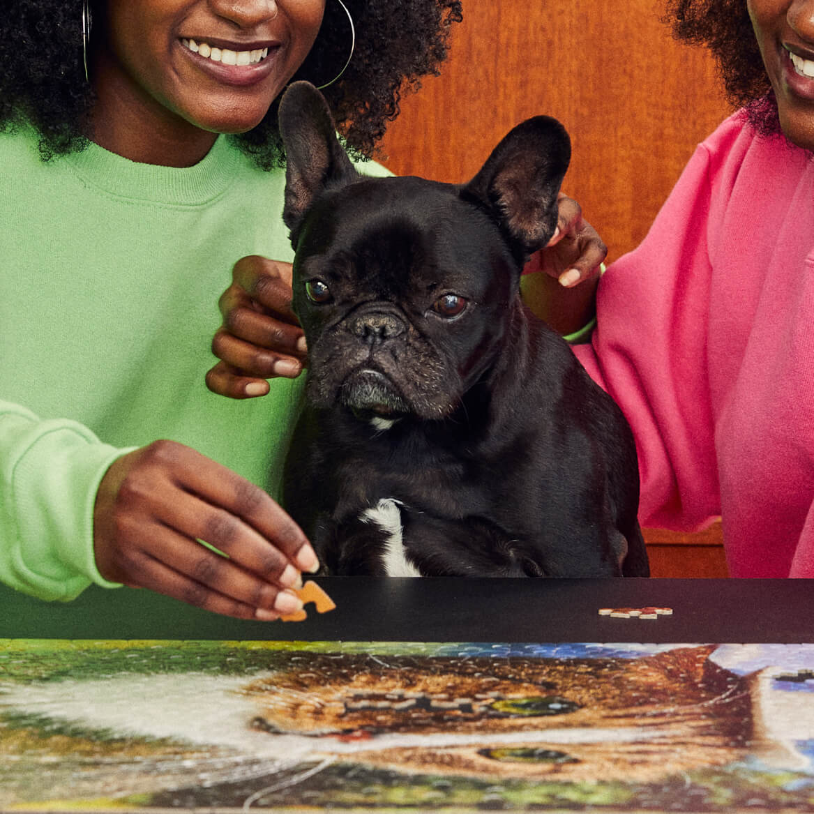 Two women smiling with a black French Bulldog while assembling a Le Puzz jigsaw puzzle