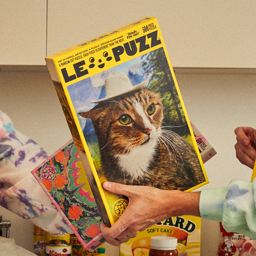 Hands holding Le Puzz Howdy Paw-tner 500-piece puzzle box featuring a cat wearing a cowboy hat