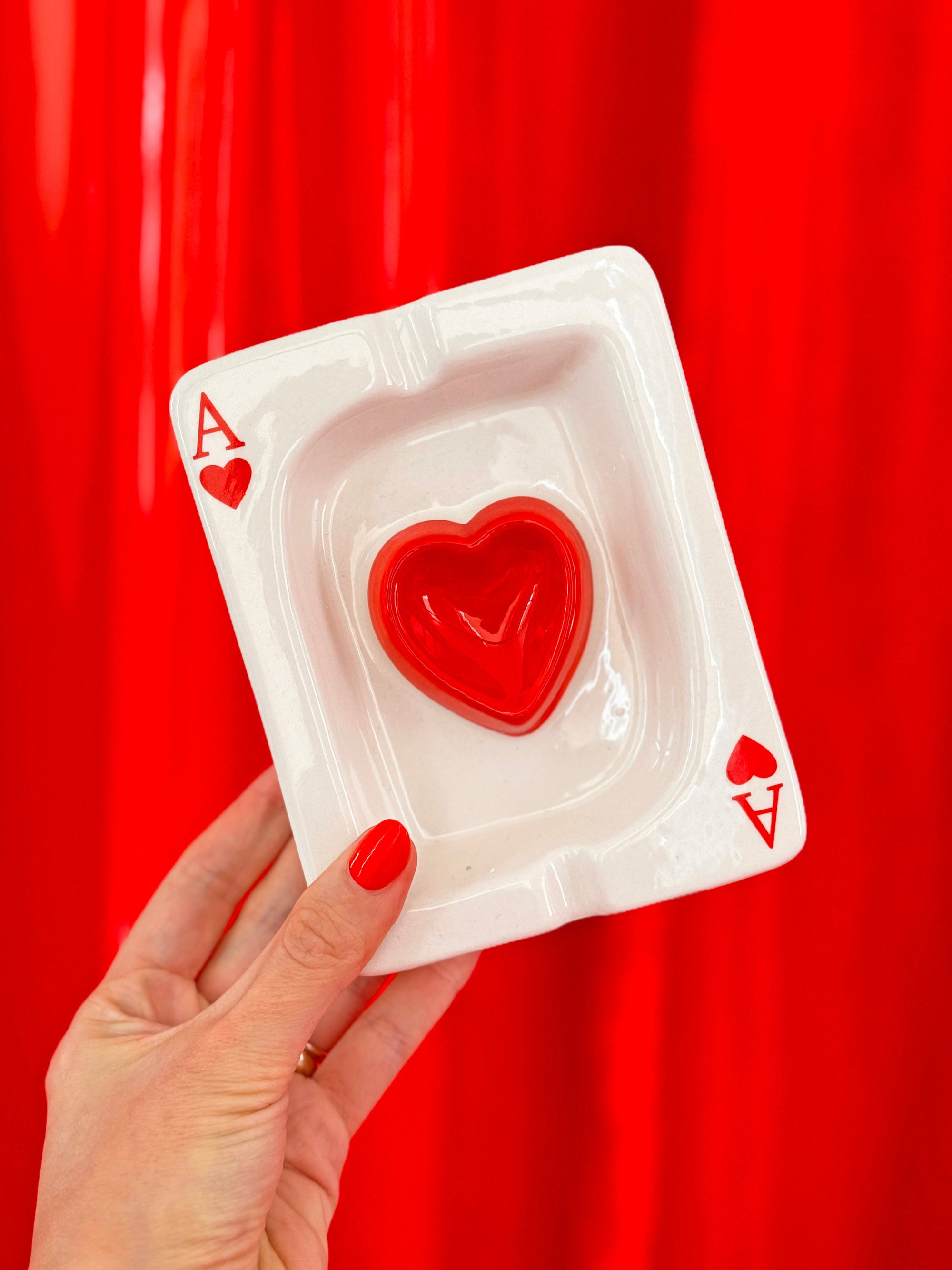 Hand holding clear acrylic ash tray shaped like Ace of Hearts playing card with red heart design