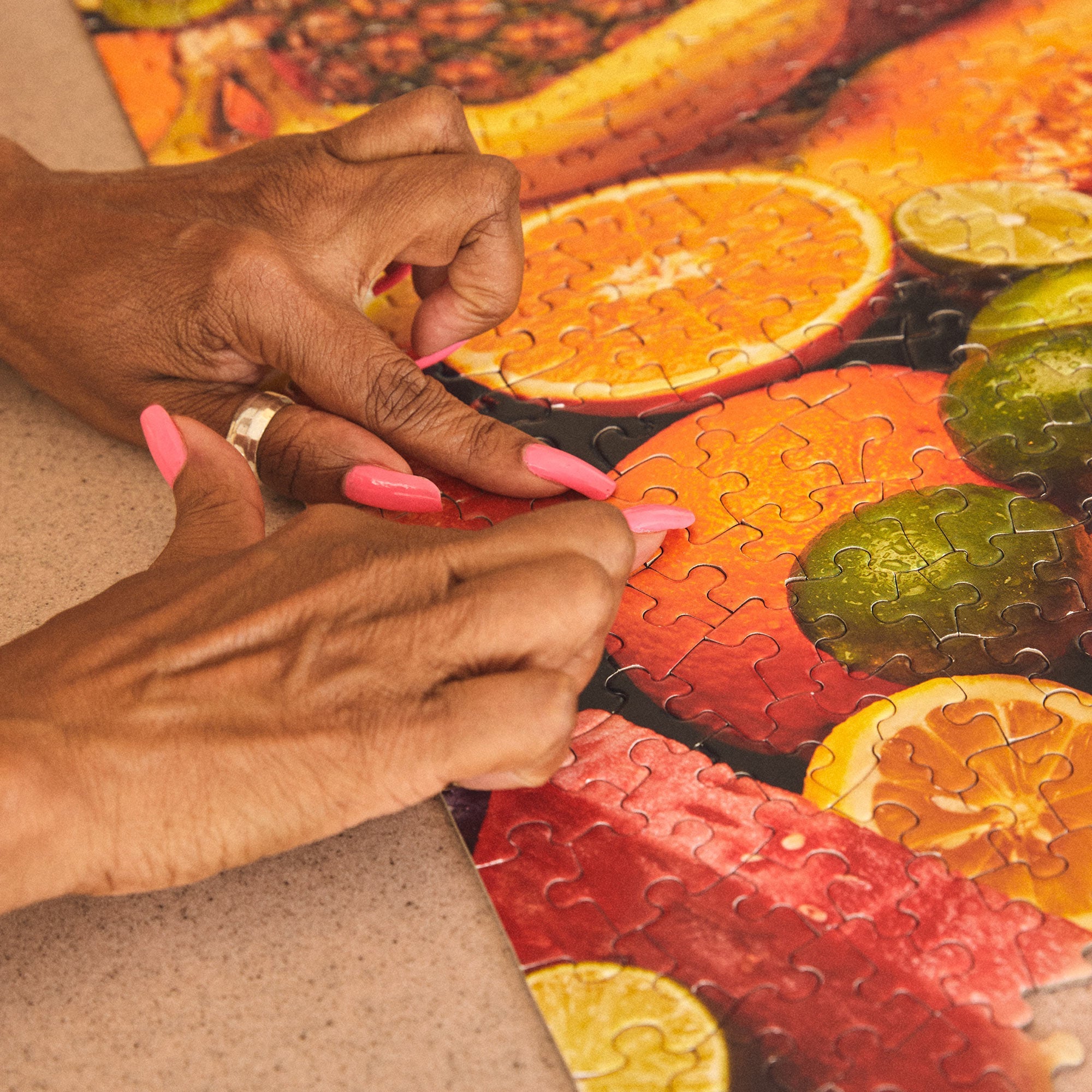 Hands with pink nails assembling Le Puzz 'Juicy' fruit-themed jigsaw puzzle on table