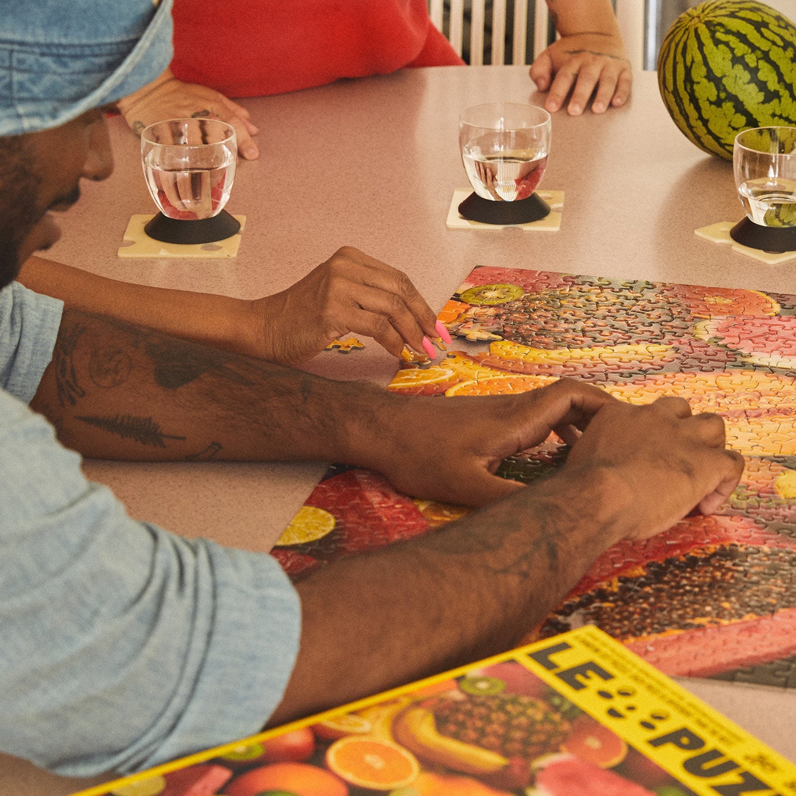 People assembling the Le Puzz Juicy fruit-themed jigsaw puzzle at a table with water glasses