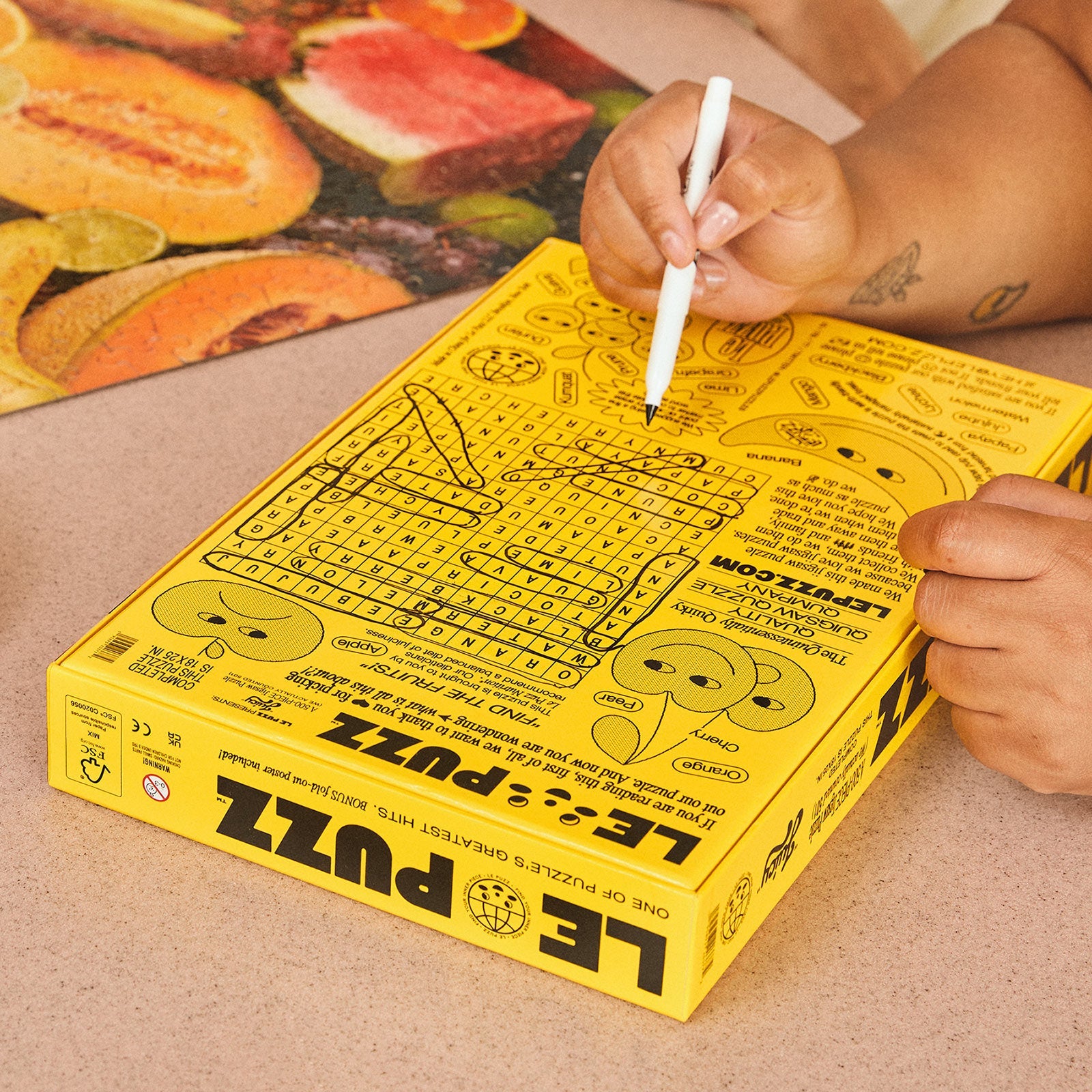Person completing word search on back of Le Puzz Juicy fruit jigsaw puzzle yellow box