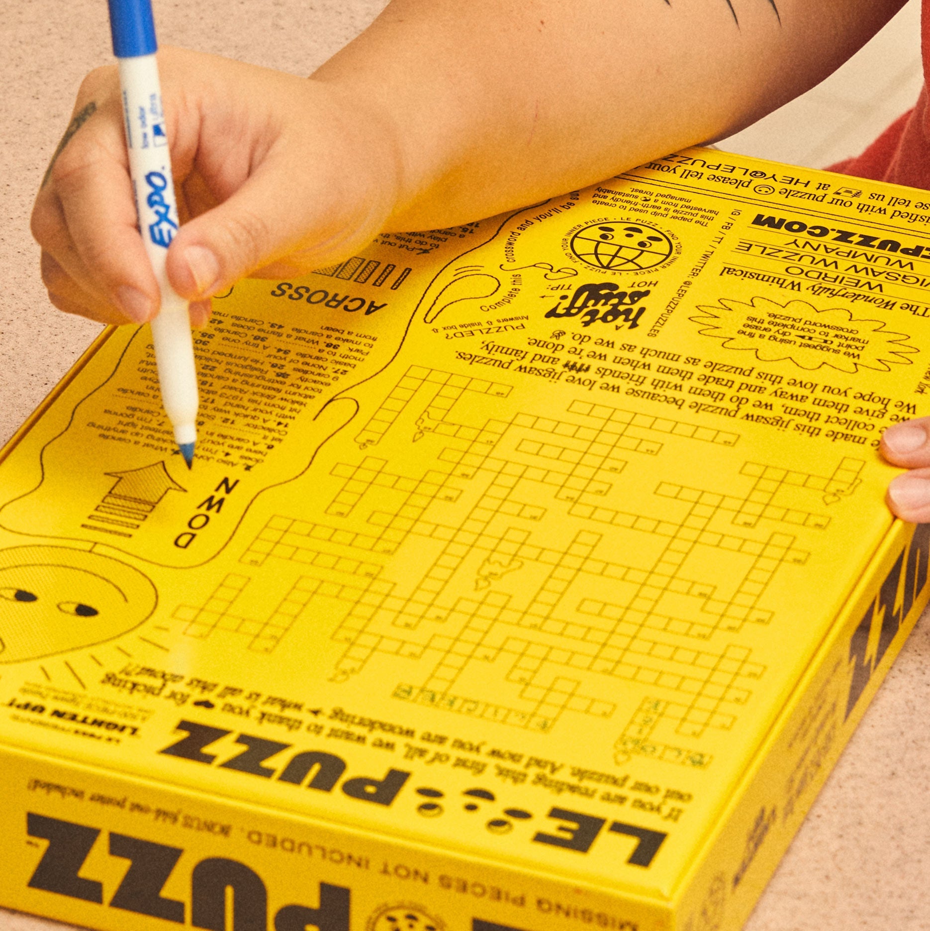Person writing on yellow Le Puzz Lighten Up jigsaw puzzle box with blue Expo marker