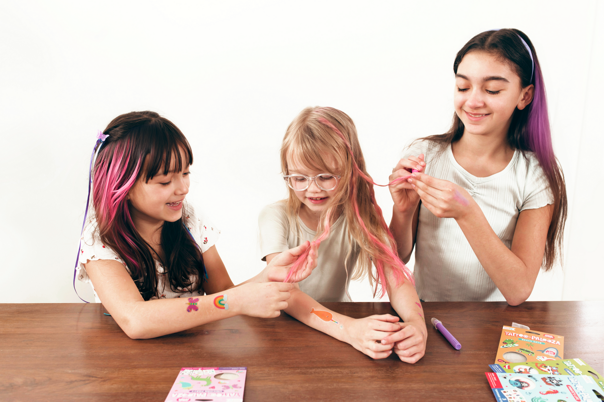 Three girls using colorful hair crayons to add pink streaks to their hair at table
