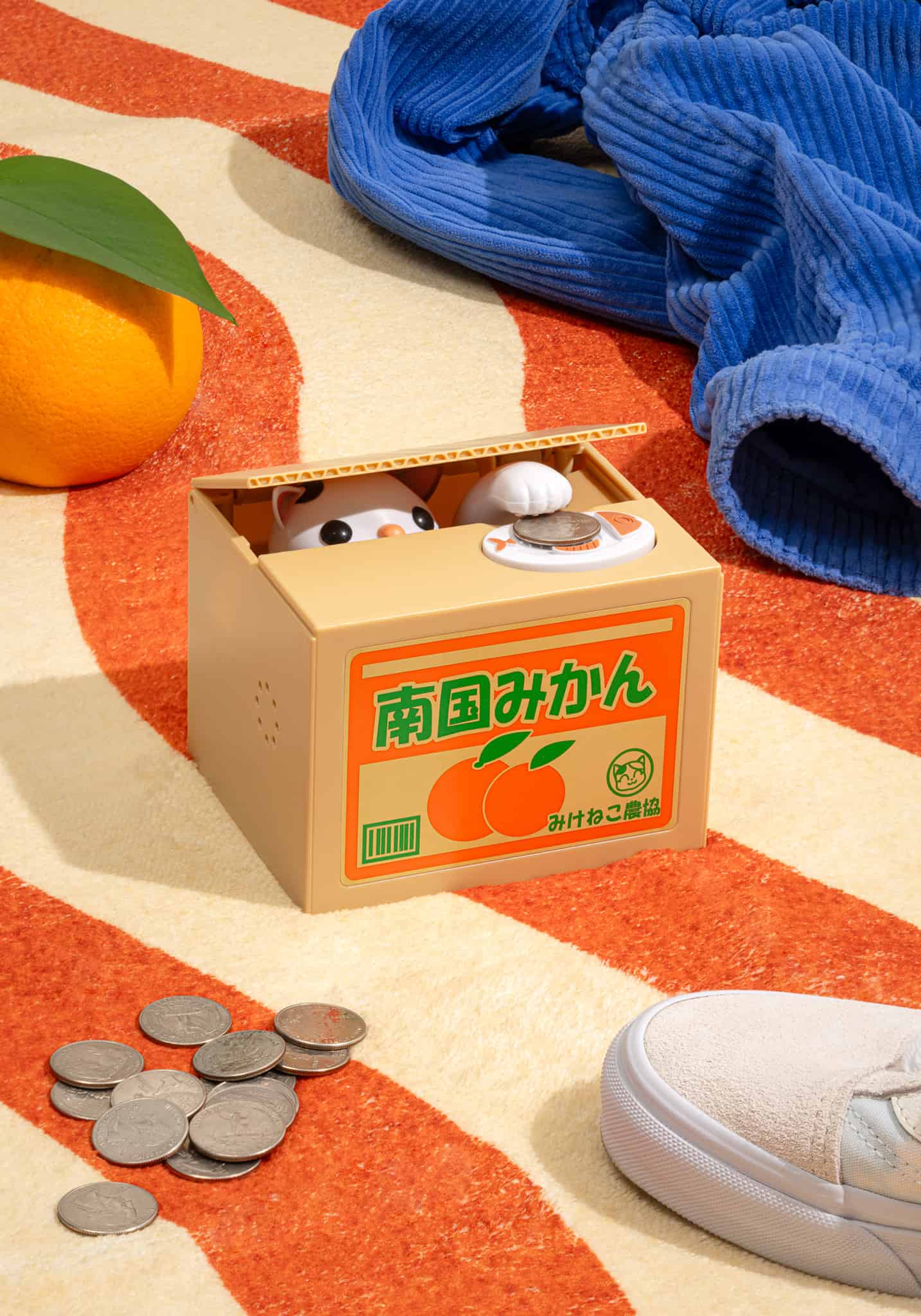Cute cat coin bank in mikan orange box with Japanese text, coins scattered nearby