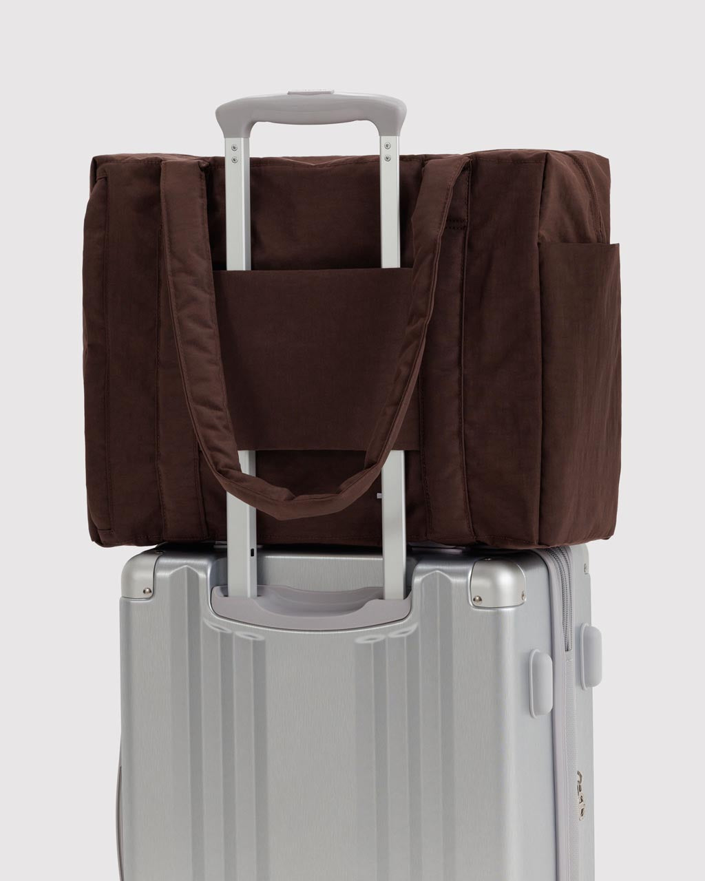 Dark brown Baggu Cloud Carry-On tote bag resting on silver hardshell luggage trolley handle