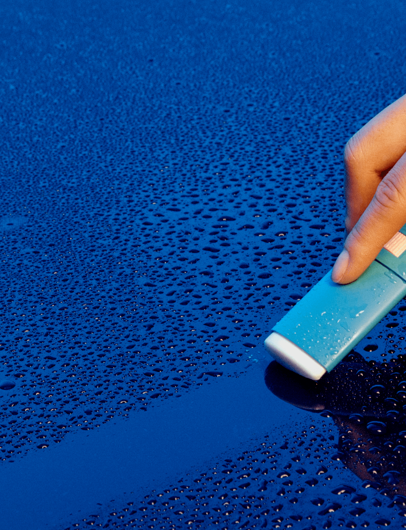 Hand holding teal Vacation Super Stick SPF 45 sunscreen on wet blue surface with water droplets