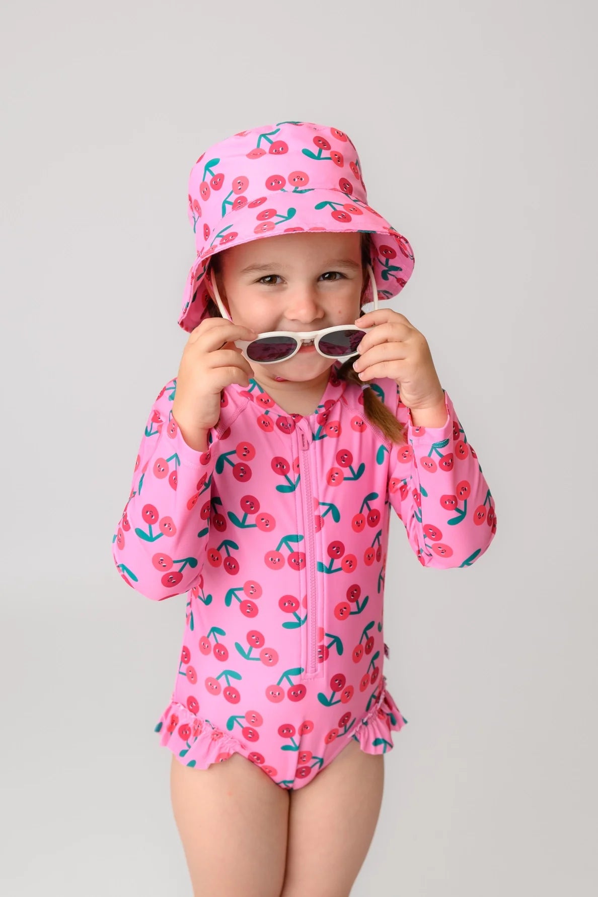 Young girl wearing pink cherry-print rash guard ruffled swimsuit with matching bucket hat and sunglasses