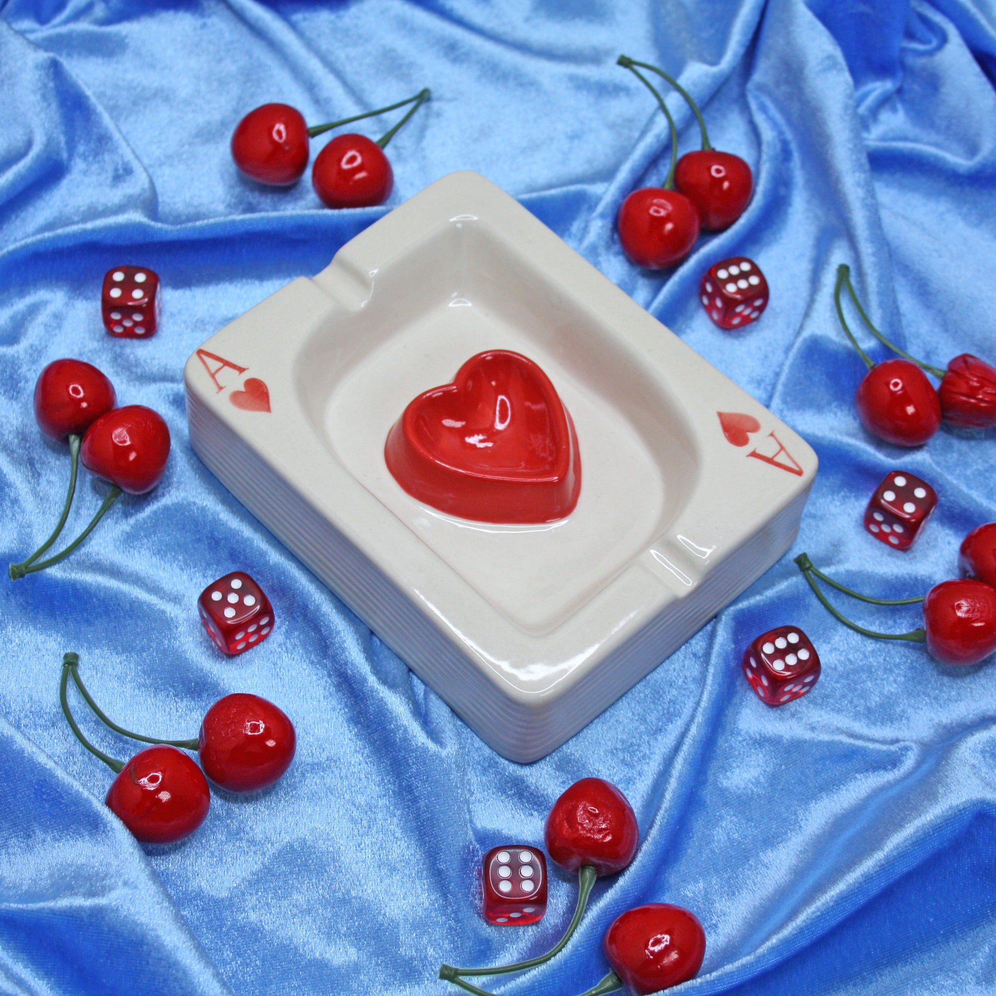 White ceramic ashtray with red ace of hearts design surrounded by cherries and dice on blue fabric