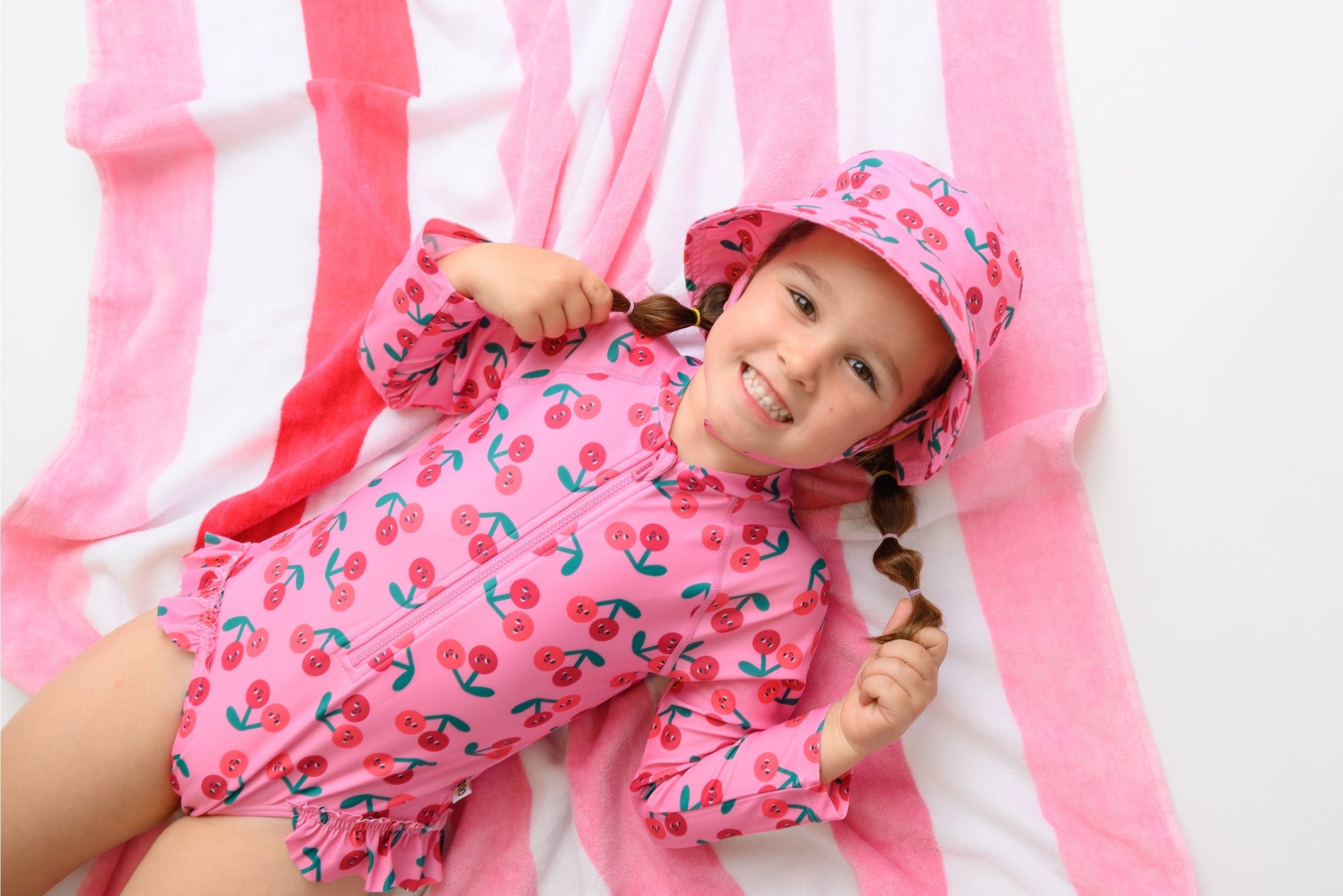 Young girl wearing pink cherry-print ruffled rash guard swimsuit and matching bucket hat lying on striped towel