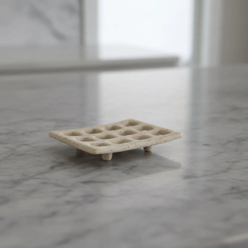 Cream colored stoneware soap dish with grid pattern and small feet on marble counter