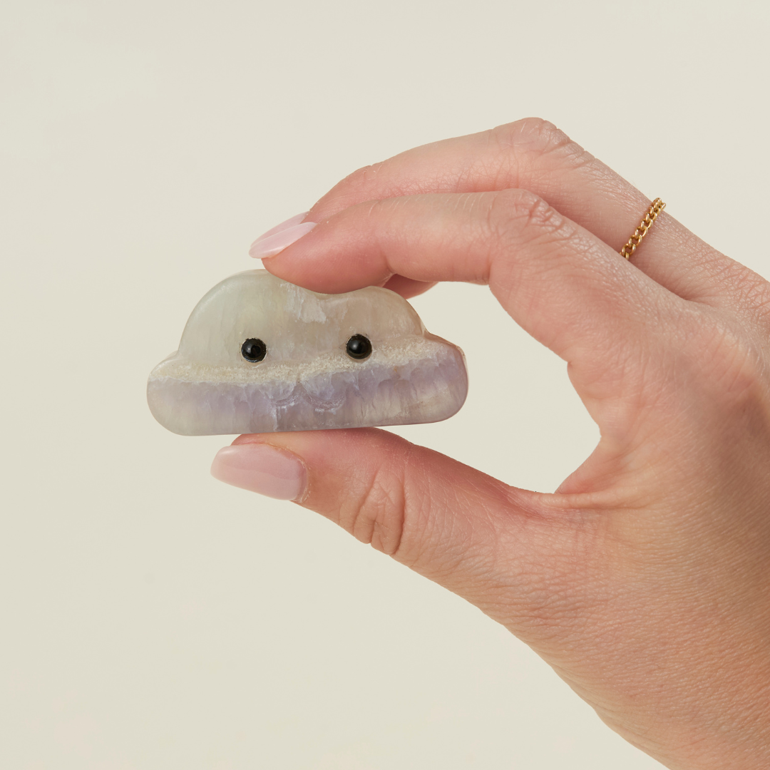 Hand holding a cloud-shaped fluorite crystal with black bead eyes against neutral background