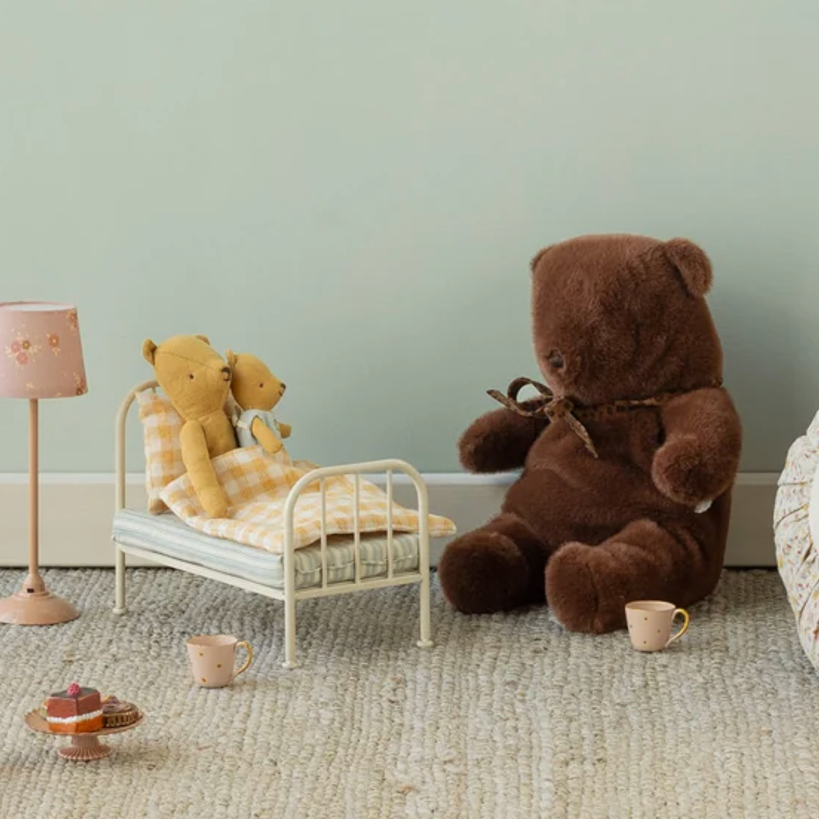 A cozy nursery-style scene with a large brown teddy bear sitting on a textured rug beside a round floral pillow, a small bed with two stuffed bears under a checkered blanket, and part of a rattan chair with a doll visible.