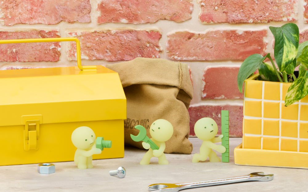 Three SMISKI glow figures holding tools beside yellow toolbox and burlap sack on brick background